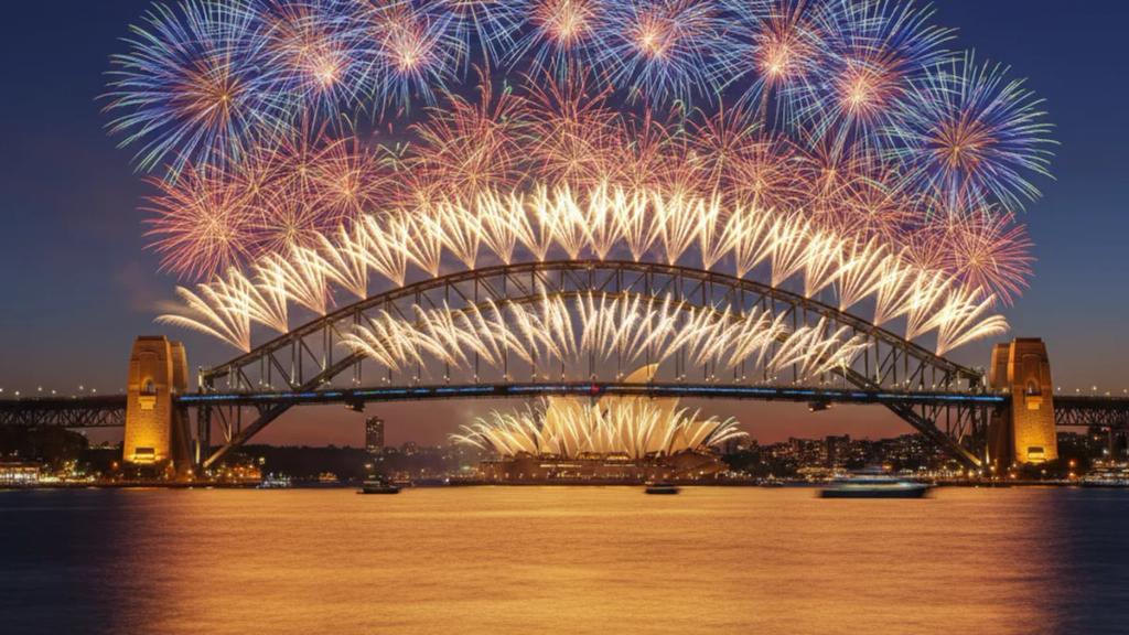 Fireworks exploding over the Sydney Harbour Bridge and Opera House at midnight on New Year's Eve, viewed from the crowded foreshore